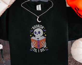 Puede incluir: Camiseta negra con un esqueleto leyendo un libro. El texto de la camiseta dice "Bookish Til I Die".
