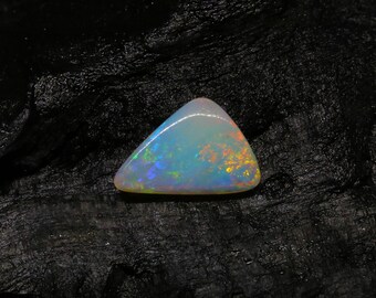 May include: A triangular opal gemstone with a pearly white base and iridescent flashes of blue, green, and orange. The polished stone has a smooth surface and is set against a dark, textured background.