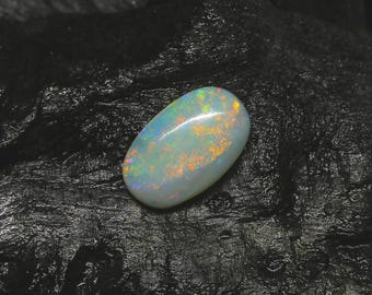 May include: A polished, oval-shaped opal gemstone. The opal displays a milky white base color with iridescent flashes of orange, yellow, and blue. The gemstone is set against a dark, textured background, highlighting its vibrant colors and unique patterns.