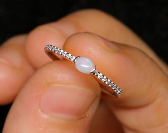 Pode incluir: Um anel de prata com uma pequena pedra de opala oval no centro. A banda é incrustada com pequenas pedras preciosas transparentes. O anel é segurado em uma mão contra um fundo escuro.