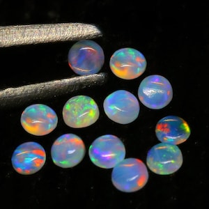 Könnte beinhalten: Eine Sammlung von 11 regenbogenfarbenen Opal-Cabochons, jeder mit einem einzigartigen Farbmuster. Die Opale sind rund und haben eine glatte, polierte Oberfläche. Sie sind in einer lockeren Gruppe auf einem schwarzen Hintergrund angeordnet.