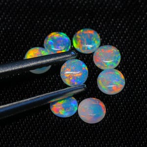 Puede incluir: Un grupo de ocho piedras preciosas redondas de ópalo iridiscente en varios tonos de azul, verde y naranja. Las piedras preciosas están dispuestas en círculo sobre un fondo de tela negro.