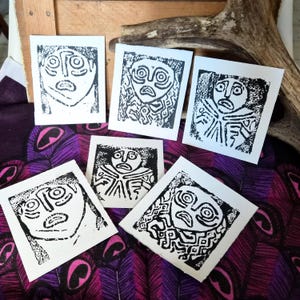 May include: A collection of six small, square art prints featuring stylized black and white faces with expressive features. The prints are arranged on a purple and pink patterned fabric, with a wooden surface and antler in the background.