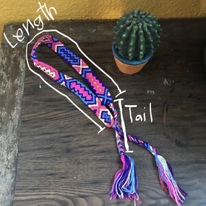 May include: A colorful friendship bracelet with a pink, blue, and white geometric pattern. The bracelet has a long tail with tassels at the end.