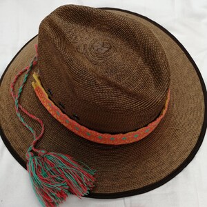 May include: A brown woven straw hat with a wide brim and a colorful braided band. The hat has a green and red tassel attached to the brim.