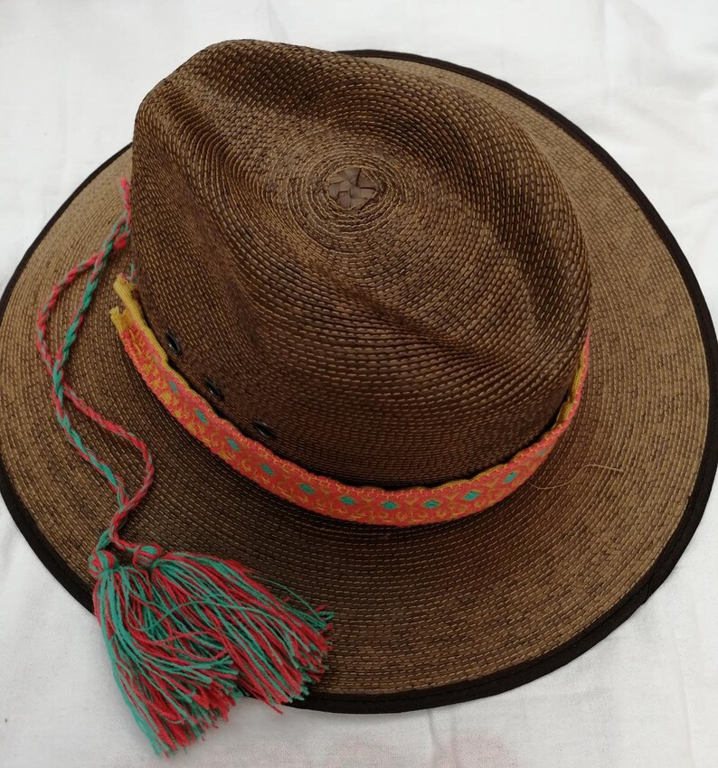 May include: A brown woven straw hat with a wide brim and a colorful braided band. The hat has a green and red tassel attached to the brim.