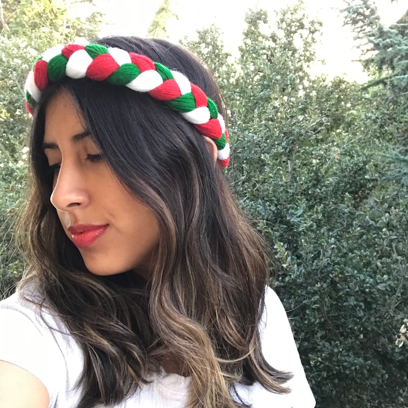 May include: A woman with long brown hair is wearing a braided headband with red, white, and green yarn. The headband is a traditional Mexican style.
