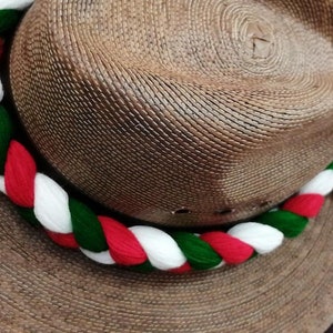 May include: A brown straw hat with a white, red, and green braided band around the crown. The hat has a wide brim and a white, red, and green tassel.