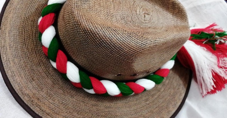 May include: A brown straw hat with a white, red, and green braided band around the crown. The hat has a wide brim and a white, red, and green tassel.