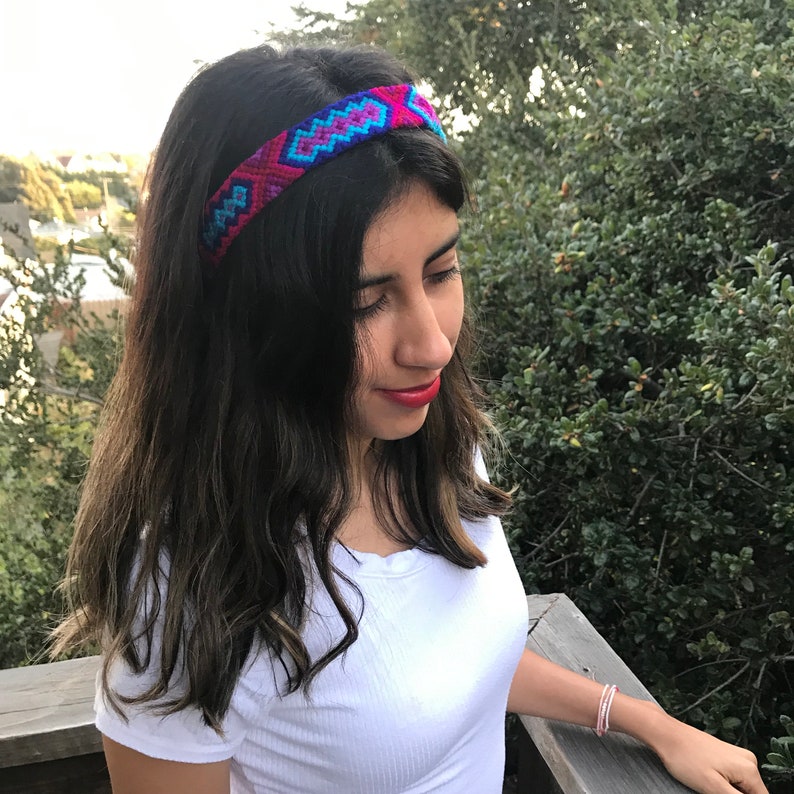 May include: A woman with long dark hair is wearing a headband with a colorful geometric pattern. The headband is made of woven fabric and has shades of pink, blue, and purple.