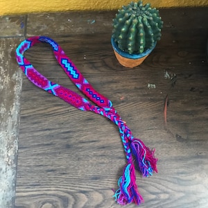 May include: A braided friendship bracelet with a pink, blue, and purple geometric pattern. The bracelet has long, fringed ends.