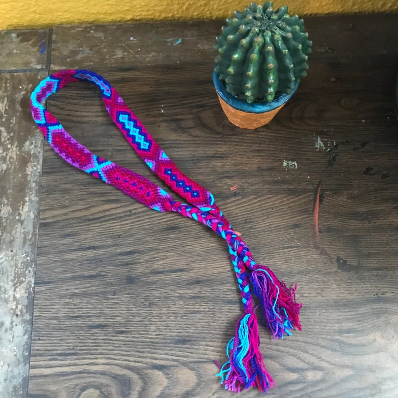 May include: A braided friendship bracelet with a pink, blue, and purple geometric pattern. The bracelet has long, fringed ends.