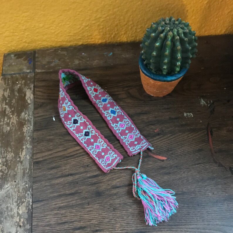 May include: A pink woven belt with a white and multi-colored geometric pattern and a long tassel. The belt is on a wooden surface.