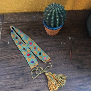 May include: A yellow and blue woven belt with colorful diamond patterns and tassels. The belt is lying on a wooden surface next to a potted cactus.