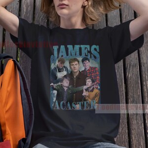 May include: Black t-shirt with a vintage-style graphic of the band "James Acaster" in blue and white text. The graphic features a photo of the band members.