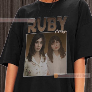 May include: Black t-shirt featuring a brown graphic of the text "RUBY Cruz" and a photograph of two women.