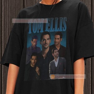 May include: Black t-shirt with a vintage-style graphic of Tom Ellis, featuring multiple images of him in different poses. The text "TOM ELLIS" is printed in blue above the images.