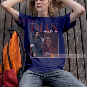 May include: A navy blue t-shirt featuring a photograph of Iman Vellani, the actress who plays Ms. Marvel, in a red and blue costume. The text "Iman Vellani" is printed in red on the shirt.