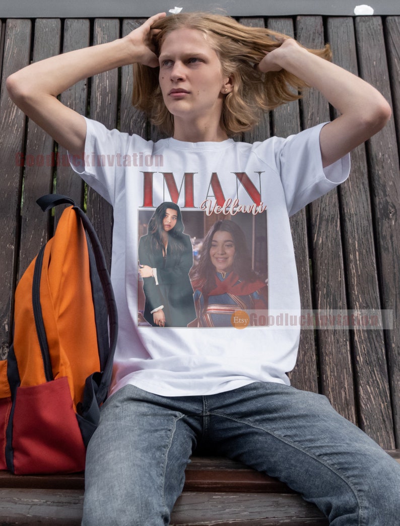May include: A white t-shirt featuring a graphic print of two women. The text "IMAN Vellani" is printed on the shirt. The women are wearing superhero costumes and are standing in front of a city skyline.