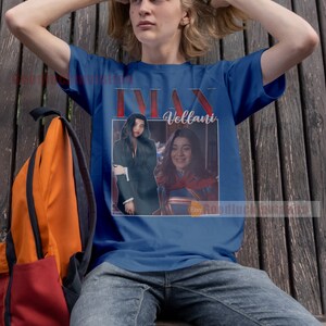 May include: A blue t-shirt featuring a photograph of a woman in a superhero costume with the text "Ms. Marvel" and "Vellani" printed on it.