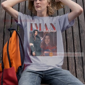 May include: A grey t-shirt featuring a graphic print of Iman Vellani as Ms. Marvel. The graphic is in a retro style with a red, white and blue colour scheme. The text "Iman Vellani" is printed in white letters on the shirt.