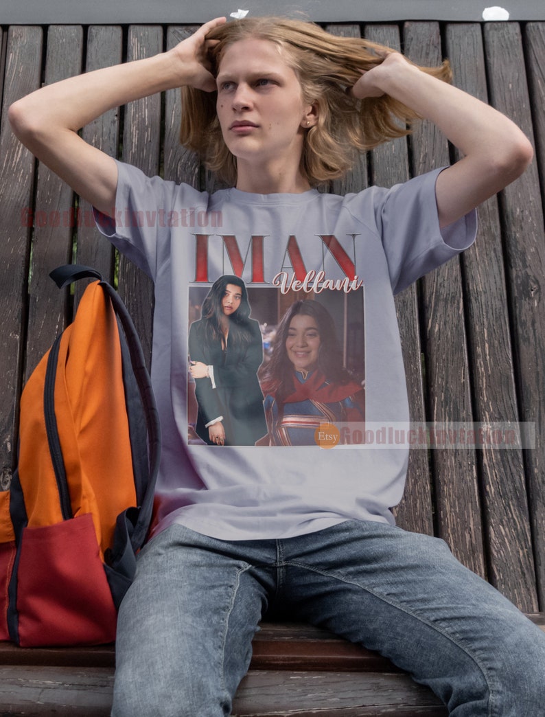 May include: A grey t-shirt featuring a graphic print of Iman Vellani as Ms. Marvel. The graphic is in a retro style with a red, white and blue colour scheme. The text "Iman Vellani" is printed in white letters on the shirt.