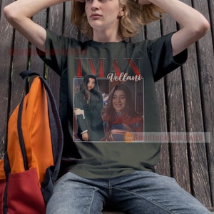May include: A dark green t-shirt featuring a vintage-style graphic of Iman Vellani, the actress who plays Ms. Marvel, in a red and blue superhero costume. The graphic also includes a smaller image of Vellani in a black suit.