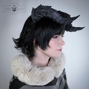 May include: A person wearing a black wig with a pair of black feathered horns on top of their head. The person is wearing a white fur collar and a gray and black striped sweater.