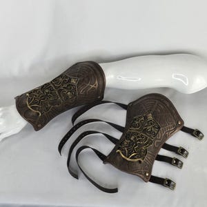 May include: A pair of brown leather arm guards with gold accents and intricate designs. The guards have straps and buckles for fastening.