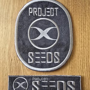 Puede incluir: Dos parches grises bordados con costuras blancas. El parche más grande tiene forma ovalada con las palabras "PROJECT SEEDS" y un logotipo circular. El parche más pequeño es rectangular y presenta el mismo texto y logotipo.
