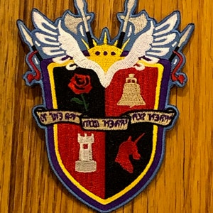 Peut inclure: Un écusson brodé coloré avec un design de bouclier. Le bouclier est divisé en quatre sections, chacune avec un symbole différent : une rose, une cloche, une tour et une licorne. Le bouclier est entouré d'une bordure bleue avec une garniture jaune. Le patch a une bannière avec le texte "Of the Gay" dans une police cursive.