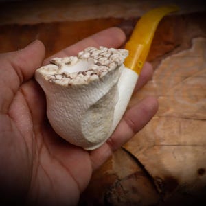 Puede incluir: Una pipa de tabaco artesanal con un cuenco blanco y una boquilla amarilla. El cuenco tiene un diseño texturizado y orgánico, y la boquilla es curva. La pipa se sostiene en una mano, mostrando su tamaño y detalle. Un accesorio para fumar único.