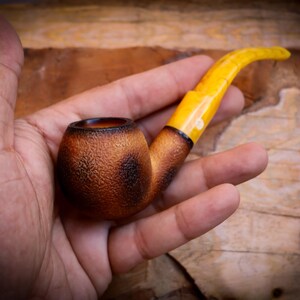 Puede incluir: Una pipa de madera marrón y negra con una boquilla de ámbar amarillo. La pipa se sostiene en una mano.
