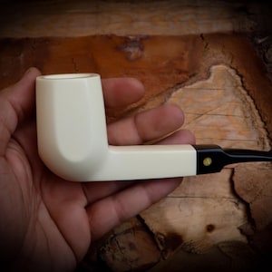 May include: Ivory-colored tobacco pipe with a black mouthpiece and a small gold-colored accent. The pipe is held in a hand, showcasing its smooth, clean design. The pipe's shape is classic, with a rounded bowl and a straight stem.