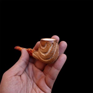 Puede incluir: Una mano sostiene una pequeña pipa de madera tallada de forma intrincada. La pipa es marrón con una cazoleta marrón claro y una boquilla roja y naranja.