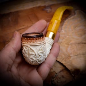 Puede incluir: Una pipa de tabaco tallada con un cuenco de color crema con diseños florales. La boquilla es de color amarillo translúcido con una banda dorada. La pipa se sostiene en una mano, mostrando los intrincados detalles de la pipa.