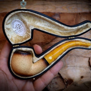 Puede incluir: Una pipa de ámbar vintage en una caja de cuero negro con ribete dorado. La caja está forrada con una tela de color crema. La pipa está hecha de ámbar y tiene una cazoleta marrón. La caja tiene una etiqueta que dice "ASTAS 1910 MERCHANDISE TEL. 1004 EMBROIDERY TURKEY".