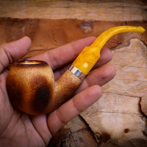 Puede incluir: Una pipa de madera marrón y negra con una boquilla de ámbar amarillo. La pipa se sostiene en una mano.