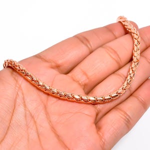 Puede incluir: Una pulsera de cadena de oro rosa con un diseño tejido. La pulsera se sostiene en una mano sobre un fondo blanco. La cadena está hecha de pequeños eslabones entrelazados.