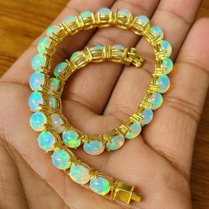 Puede incluir: Una pulsera dorada con una serie de ópalos ovalados. Los ópalos tienen una base blanca lechosa con destellos azules y verdes. La pulsera se sostiene en una mano, mostrando el diseño y el color de la joya.