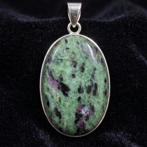 Puede incluir: Un colgante de forma ovalada con una piedra de zoisita pulida que presenta una base verde moteada, inclusiones negras oscuras y manchas de color rosa magenta vibrante. La gema está engastada en un marco de metal plateado liso.