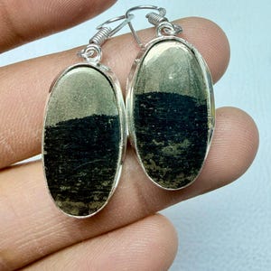 Può includere: Orecchini color argento con pietre di pirite ovali. Le pietre hanno un aspetto scuro e strutturato, incastonate in montature in metallo argentato e ganci curvi. Gli orecchini sono mostrati su sfondo chiaro.