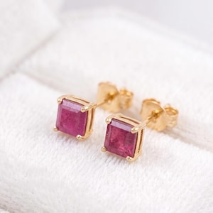 May include: A pair of stud earrings featuring square-cut, deep pinkish-red gemstones set in four-prong yellow gold settings. The earrings have standard post backs and are displayed clearly to show the faceted surface of the stones.