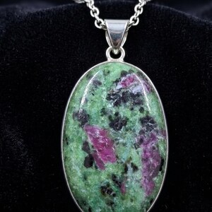 Puede incluir: Un colgante de forma ovalada con una piedra de zoisita pulida que presenta inclusiones de color verde vibrante, magenta intenso y negro. La piedra está engastada en un bisel de metal plateado liso y cuelga de una cadena de eslabones plateada.