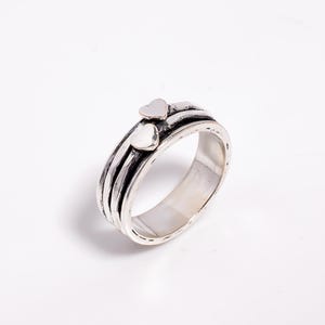 May include: A silver ring with two small heart-shaped accents. The ring has a textured band with dark grooves and a polished interior. The hearts are a lighter silver color, adding a contrasting detail to the design.