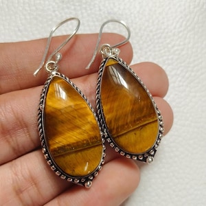 May include: A pair of teardrop-shaped earrings featuring polished tiger's eye stones. The stones are a warm brown color with golden stripes. Each earring has a silver-colored metal frame with a decorative rope-like border and a hook closure.