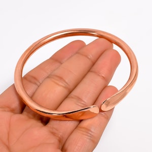 Puede incluir: Un brazalete abierto de color cobre. El brazalete tiene un acabado liso y pulido y un diseño simple y minimalista. El brazalete se sostiene en una mano, mostrando su forma y color.