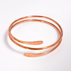May include: A copper-colored arm cuff bracelet. The bracelet wraps around the arm with two spiraling bands. The ends of the bracelet have a hammered texture. The bracelet is displayed on a white background.