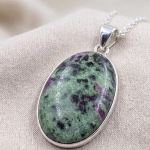 Puede incluir: Un colgante de forma ovalada con una piedra de zoisita pulida con inclusiones moteadas en verde y morado oscuro, engastado en un bisel de metal de tono plateado liso. El colgante está unido a una delicada cadena de eslabones plateada.