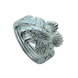 Puede incluir: Un anillo de rompecabezas plateado con un diseño detallado de águila. El águila está representada con las alas extendidas, y el anillo tiene múltiples bandas entrelazadas. El anillo está hecho de un material metálico brillante.
